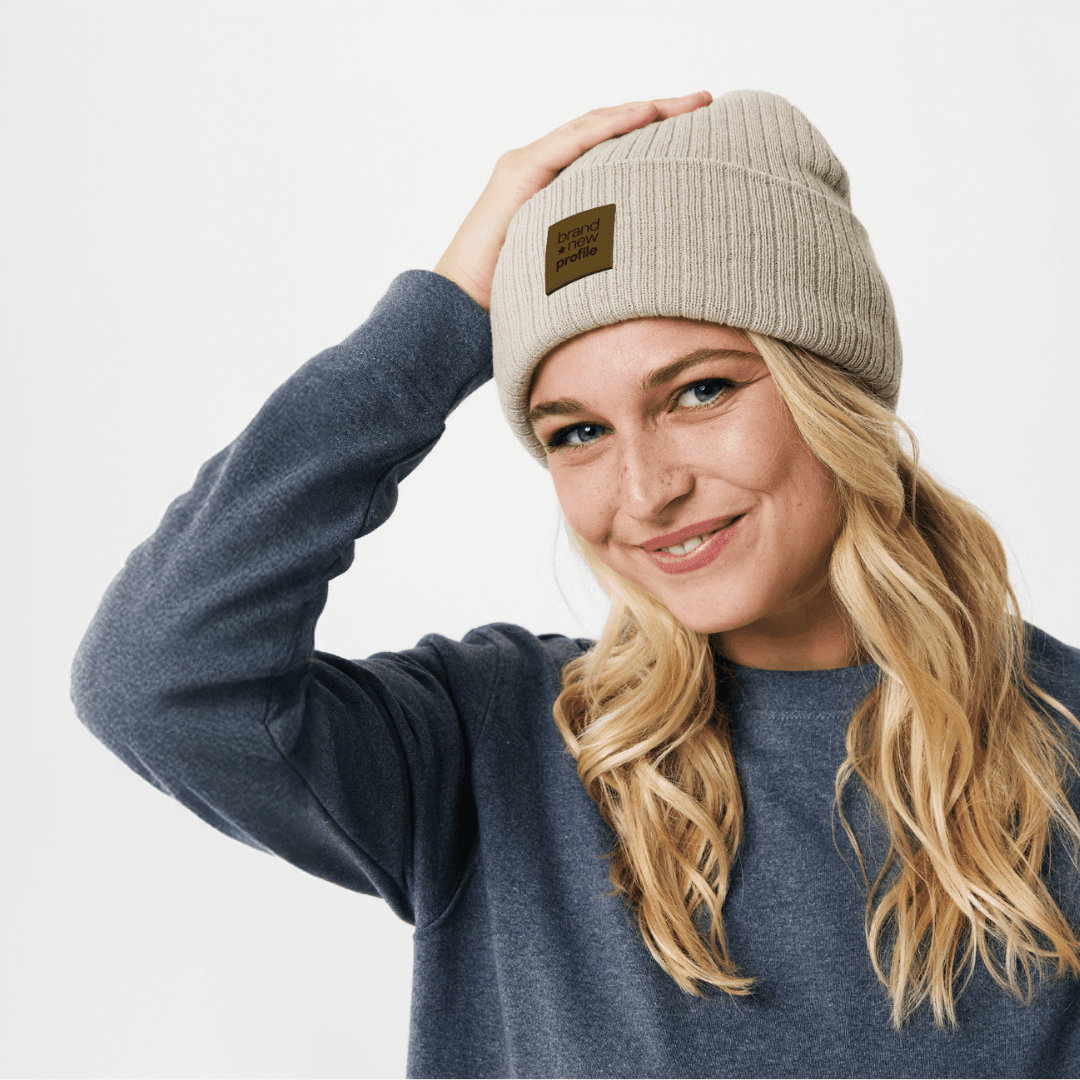 Leende kvinna som justerar en beige, ribbstickad mössa med ett lädermärke som har en egen logotyp.